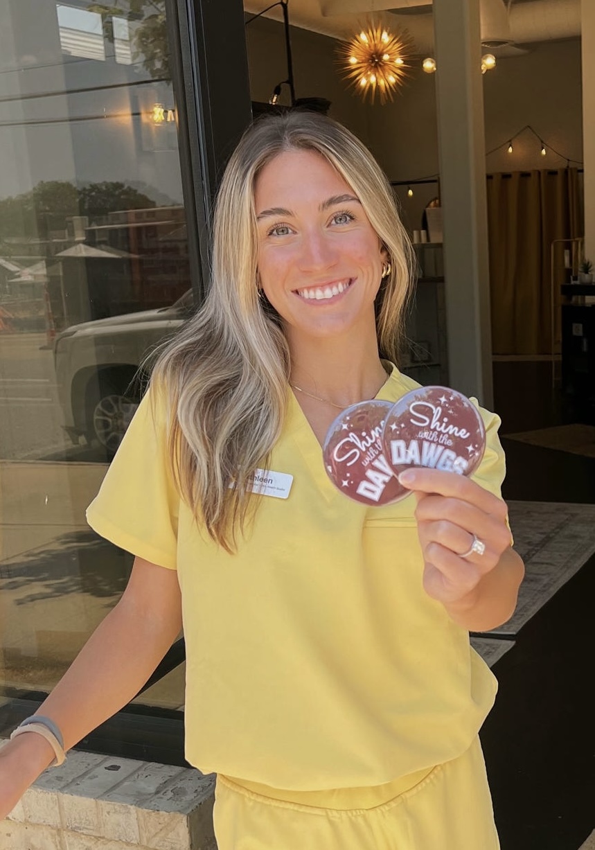 Shine Starkville esthetician Kathleen in yellow scrubs holding Shine with the Dawgs buttons
